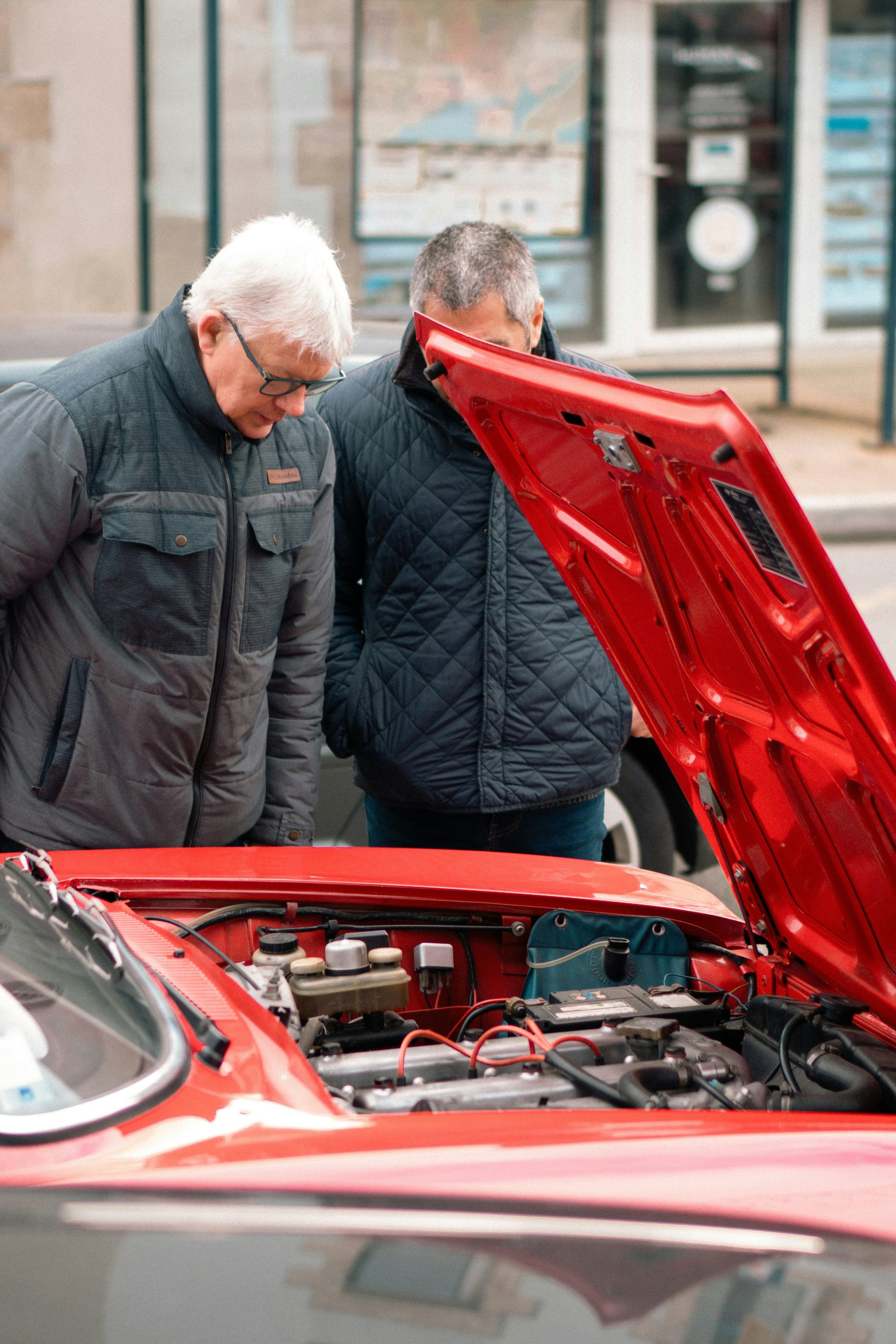 two men looking under the hood of a red car